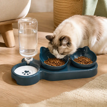 A cat eats from two bowls of dry food on a dark blue pet feeding tray while drinking water from a clear bottle, placed on a wooden floor in a cozy indoor setting.