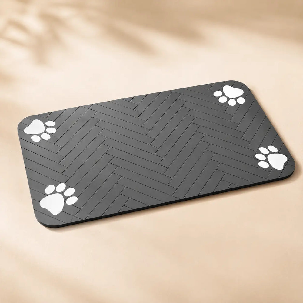 A rectangular black mat with rounded corners, featuring a dark gray herringbone pattern and four white paw print designs in the corners, shown on a beige background with soft leaf shadows.