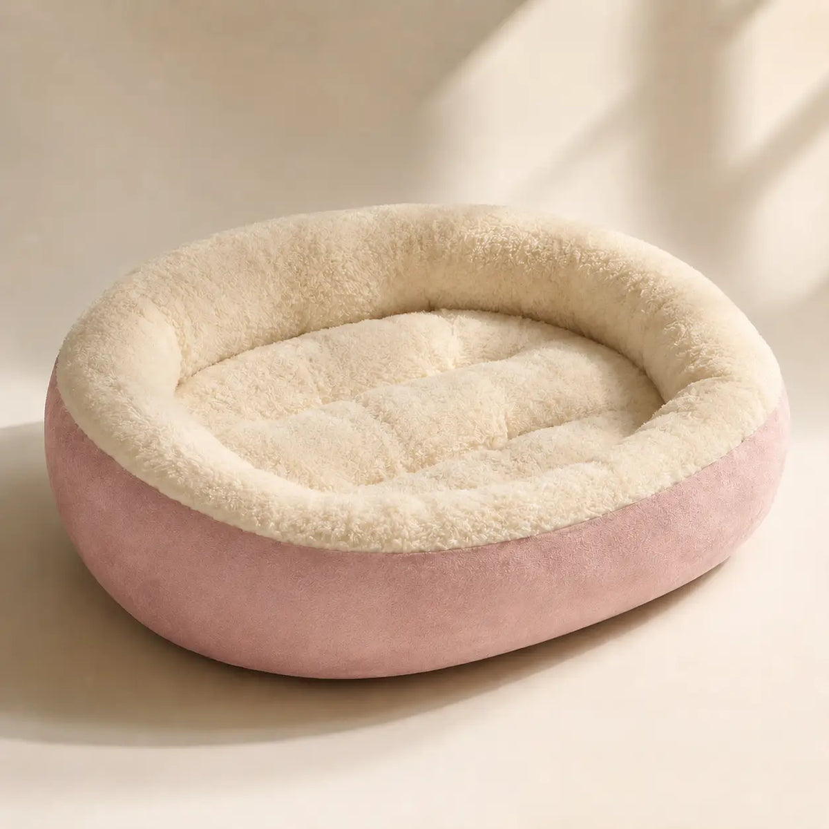 A round pet bed with a dusty pink velvet exterior and a cream sherpa fleece interior, photographed on a warm beige background with soft window-shadow lighting.