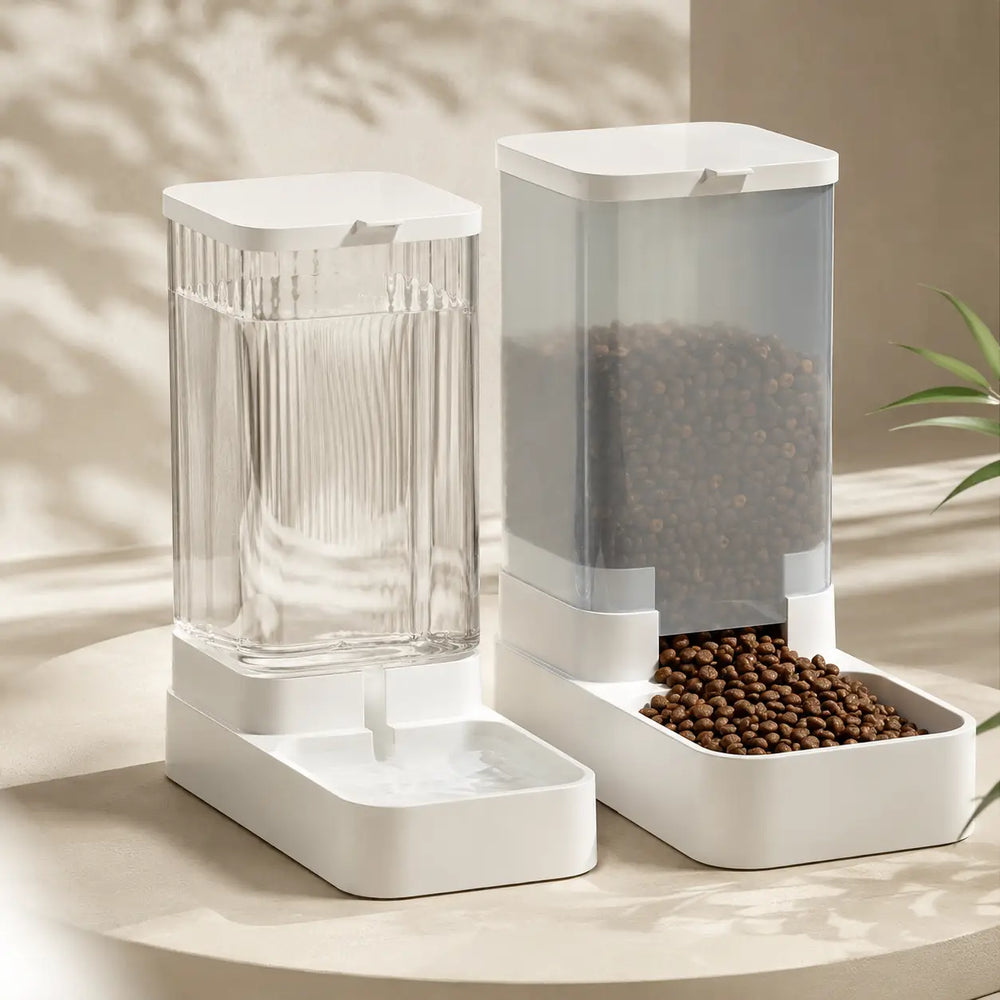 White gravity pet water dispenser and food dispenser filled with kibble, standing side by side on a beige surface in warm sunlight.