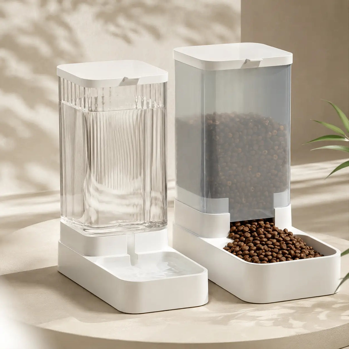 White gravity pet water dispenser and food dispenser filled with kibble, standing side by side on a beige surface in warm sunlight.