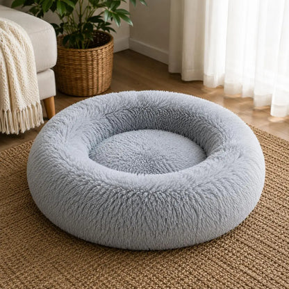Light grey plush donut pet bed with thick raised cushioned sides and a soft shaggy texture, placed on a natural woven rug beside a cream armchair and potted plant in a bright living room with sunlight coming through sheer curtains.