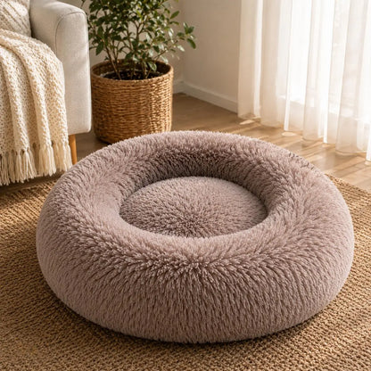Taupe plush donut pet bed with thick raised cushioned sides and a soft shaggy texture, placed on a natural woven rug beside a cream armchair and potted plant in a bright living room with sunlight coming through sheer curtains.