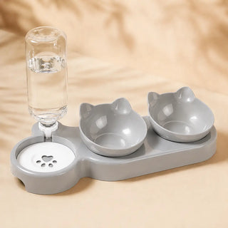 A gray pet water fountain and double food bowls set with cat-ear bowl designs and a clear water bottle, photographed on a warm beige surface.